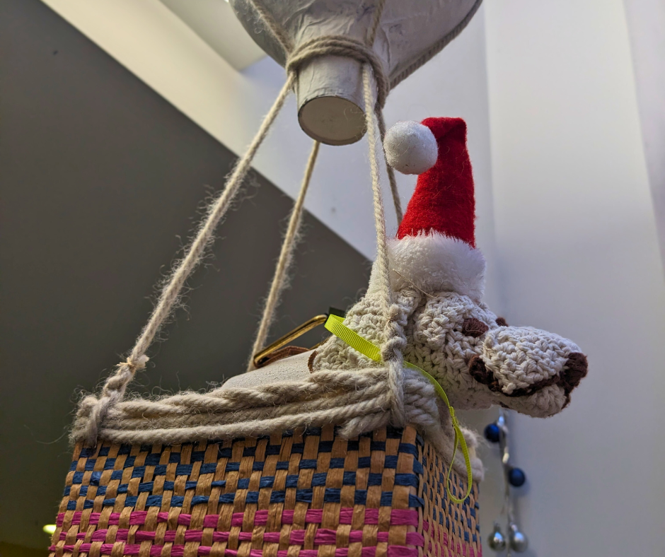 A crotchet guide dog in a Santa hat sitting in the woven basket of a mini hot air balloon.