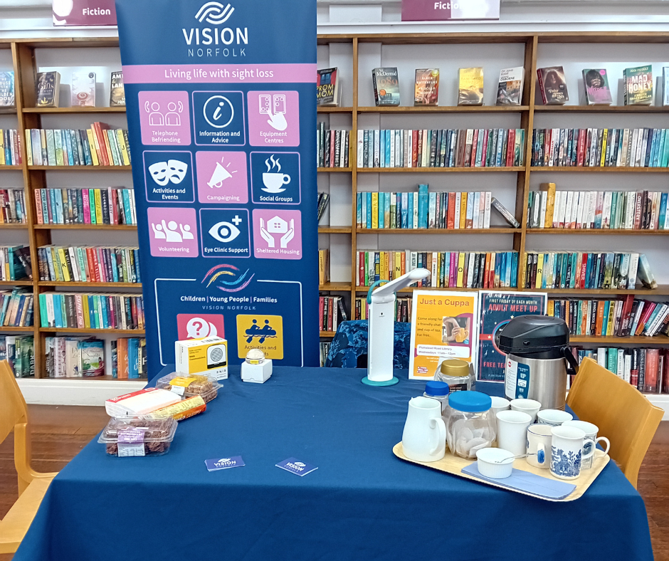 A table set up with tea, coffee, snacks and information for a Vision Norfolk drop-in cafe in the Plumstead Road Library in Norwich. There is a banner with information about Vision Norfolk in the background in front of the shelves of books.