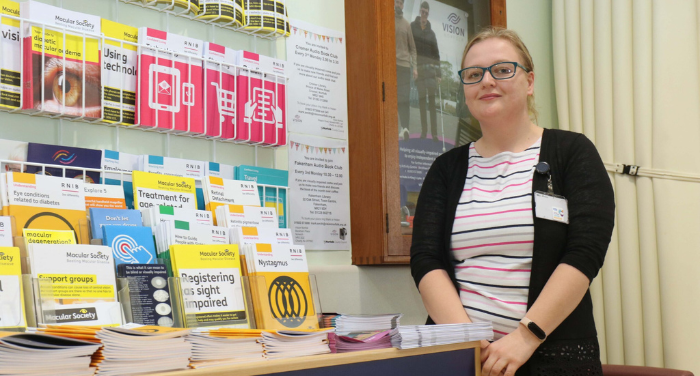 Abbi Roberts, Eye Clinic Liaison Officer in Norwich, sitting in front of a display of information for patients with sight loss.