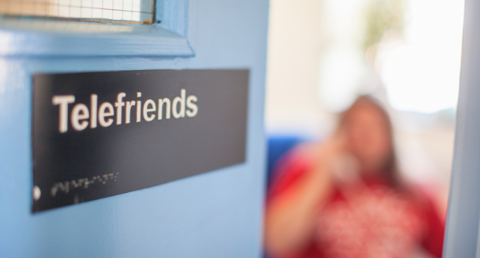A blue door with a label reading "Telefriends" in text and Braille. Blurred in the background someone is on the phone.