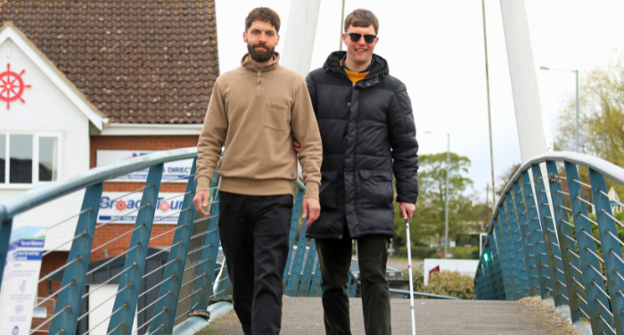 Two men walking over a bridge. The man on the right is walking with a white cane and the man on the right is guiding him.