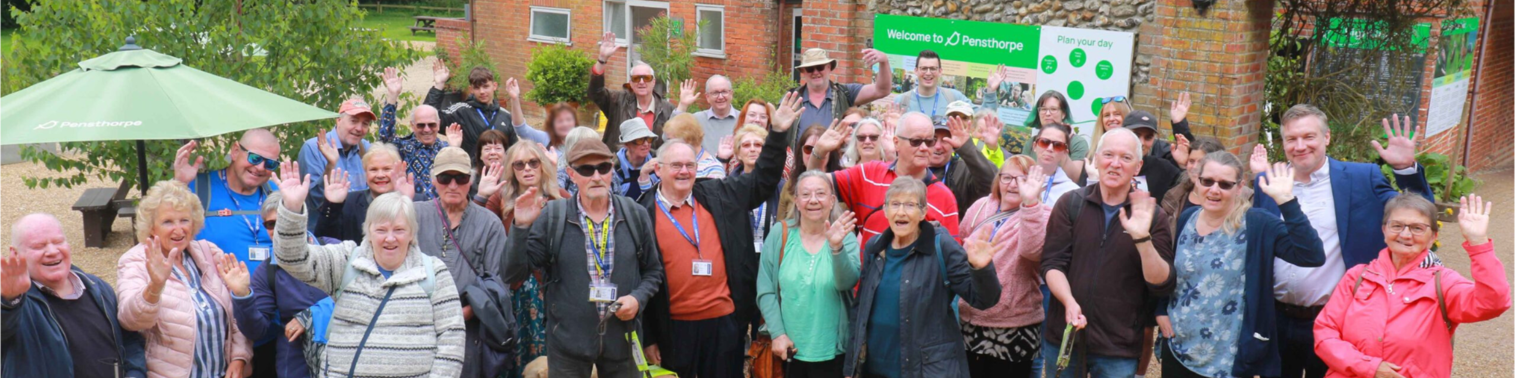 A group of volunteers, guide dogs and Vision Norfolk staff waving at the camera outside of Pensthorpe Nature Reserve in Fakenham.