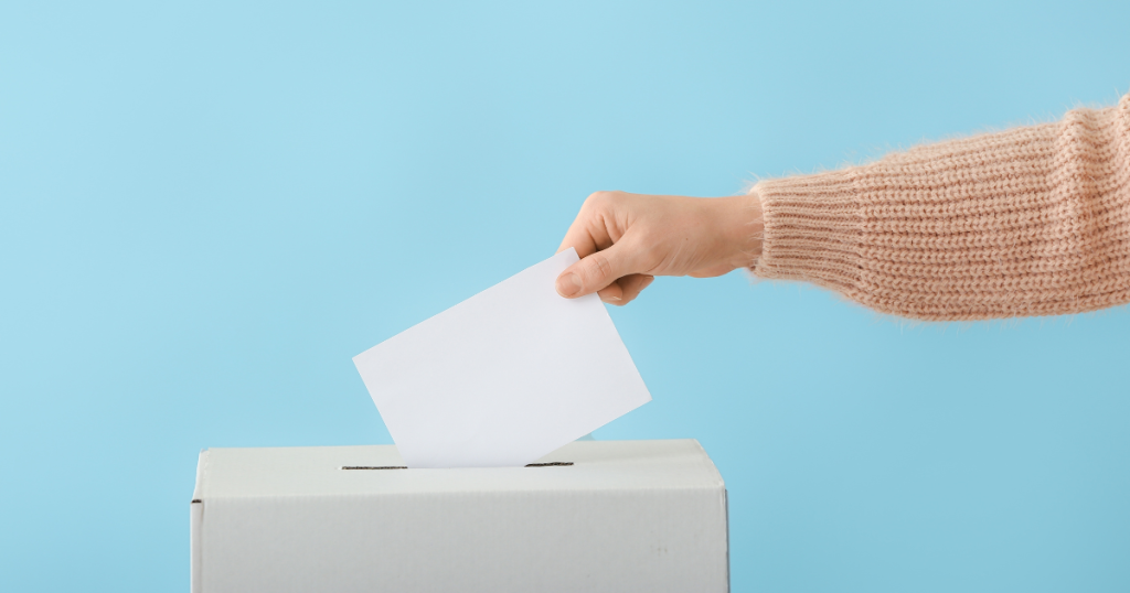 An arm wearing a pink jumper placing a slip of paper in a voting box.