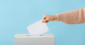 An arm wearing a pink jumper placing a slip of paper in a voting box.