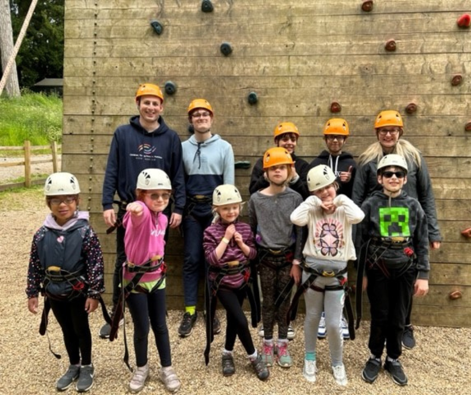 Eleven young people, Vision Norfolk staff and volunteers posing in front of an outdoor climbing wall wearing helmets and harnesses.