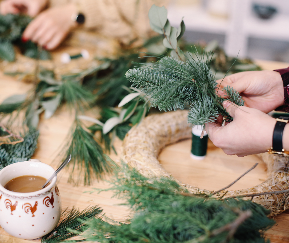 A pair of hands putting together evergreen twigs to make a wreath. There is a cup of coffee and art supplies on the table.