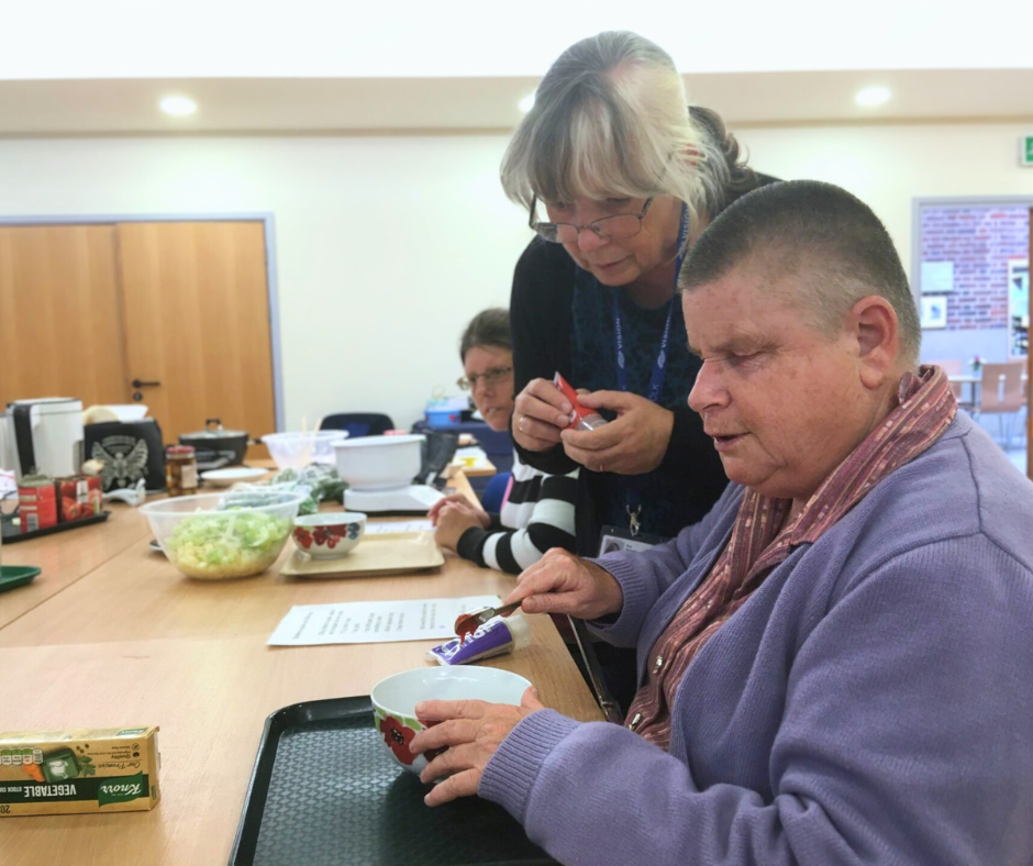 A woman in a purple cardigan is measuring tomato paste into a bowl while another woman watches over her shoulder. In the background the table is covered with bowls and various cooking instruments and there is another participant sitting in front of a tray.