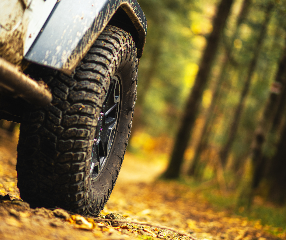 One of the font wheels of an off road vehicle with a dirt road through the woods in the background. The wheel is covered with mud.